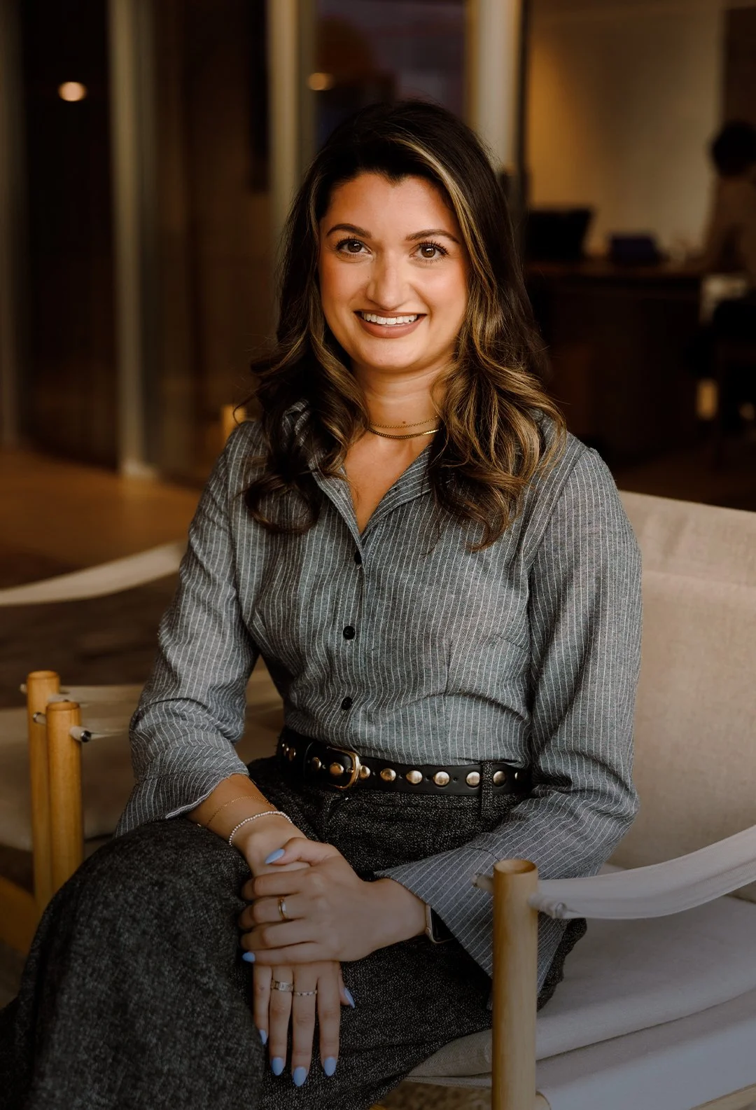 A woman with long, wavy brown hair and light skin, sitting on a beige chair in a warmly lit indoor setting, wearing a gray pinstripe button-up shirt and dark pants, smiling at the camera.