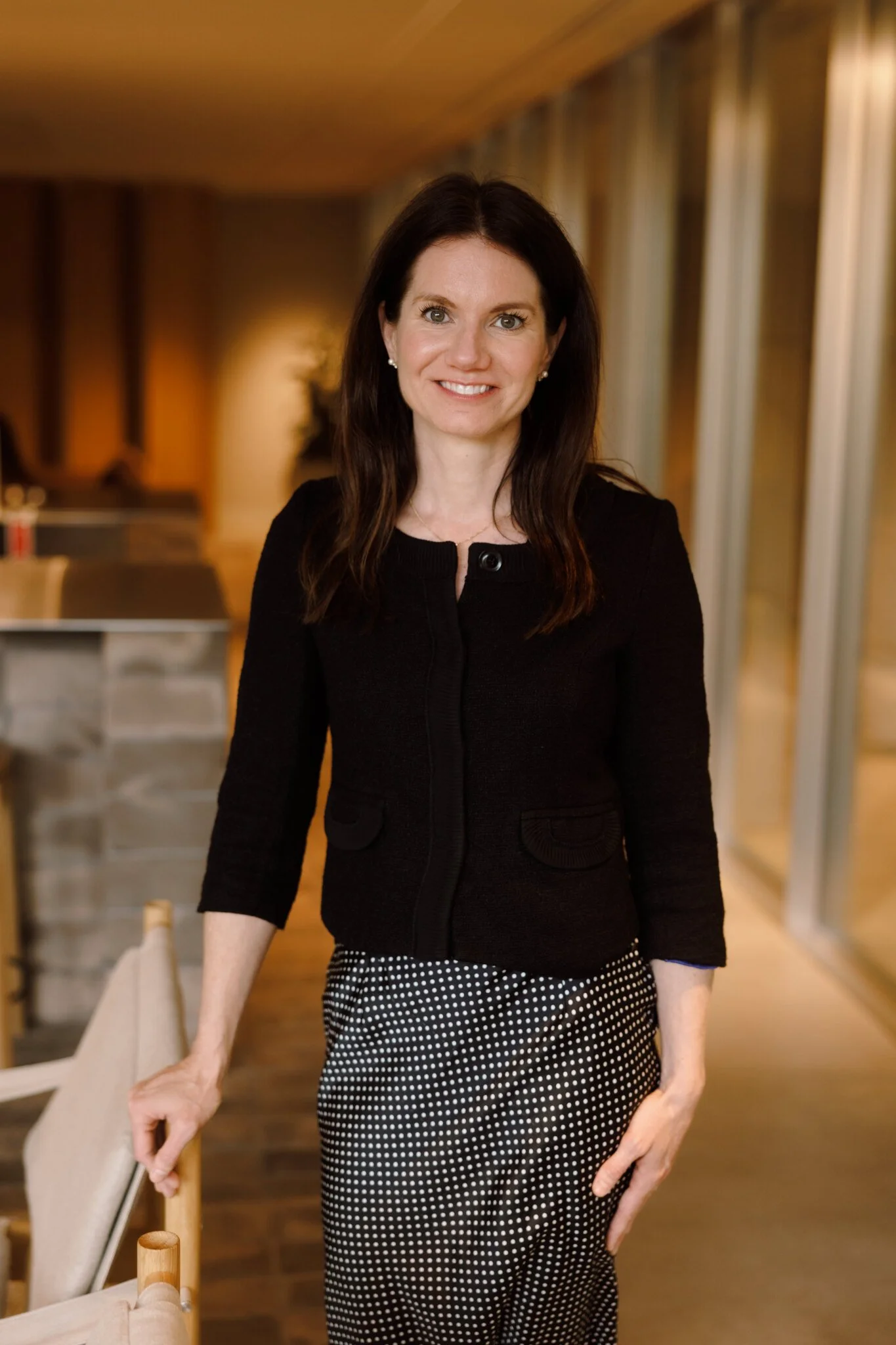 A woman with dark brown hair and a light complexion, wearing a black cardigan and black-and-white polka dot pants, standing indoors with a warm wooden background and smiling at the camera.