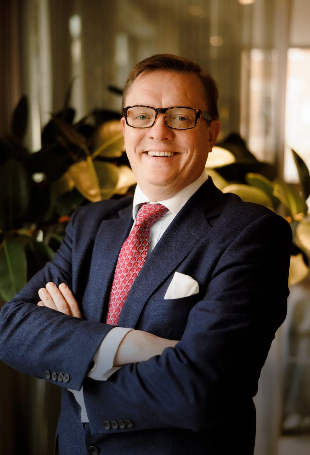 A smiling man in a navy blue suit, white shirt, and red patterned tie with arms crossed, standing in front of green plants and a window.