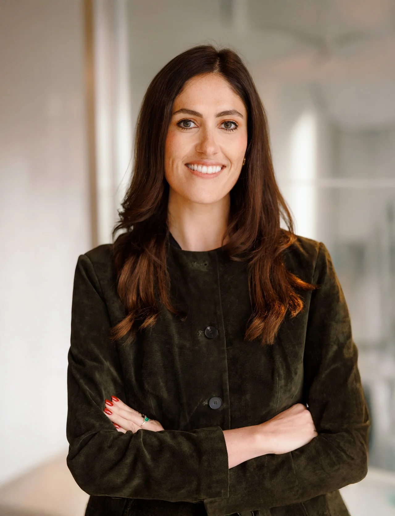A woman with long brown hair, wearing a dark button-up jacket, smiling, with her arms crossed.