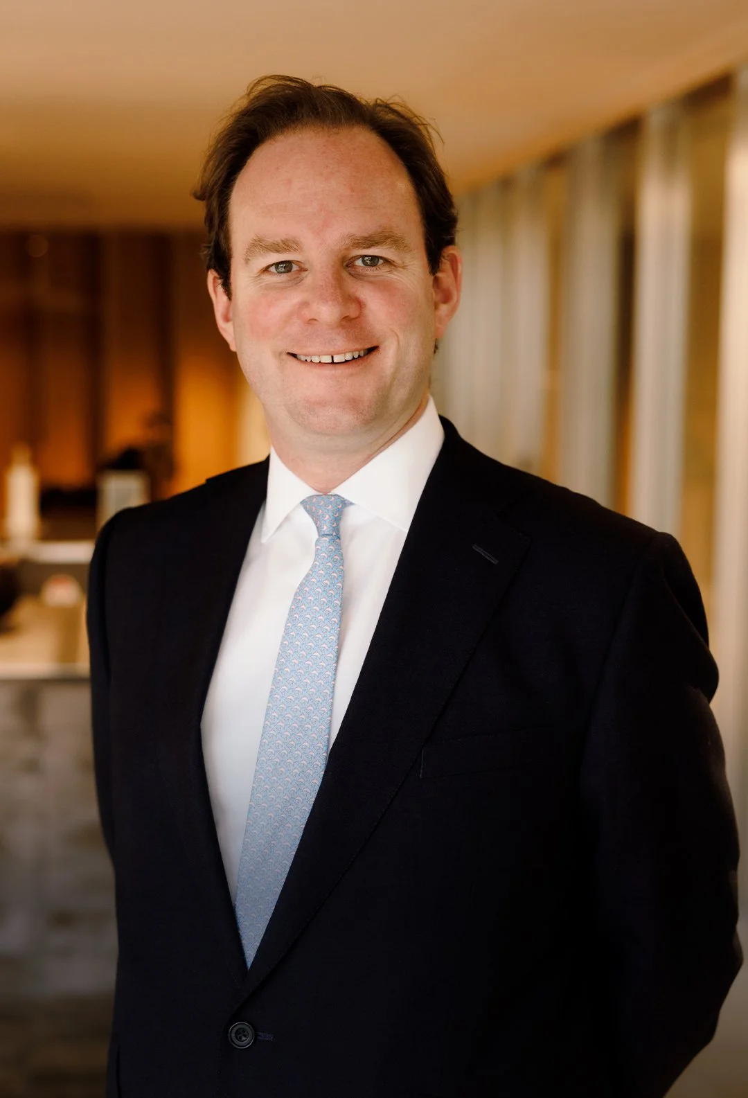 A smiling man in a black suit and light blue tie standing in a warmly lit room with curtains and furniture in the background.