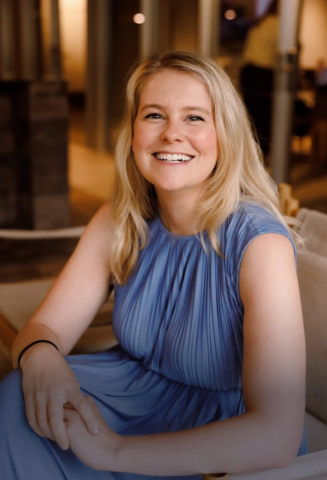 A smiling woman with blonde hair wearing a blue sleeveless dress, sitting indoors in a warmly lit environment.