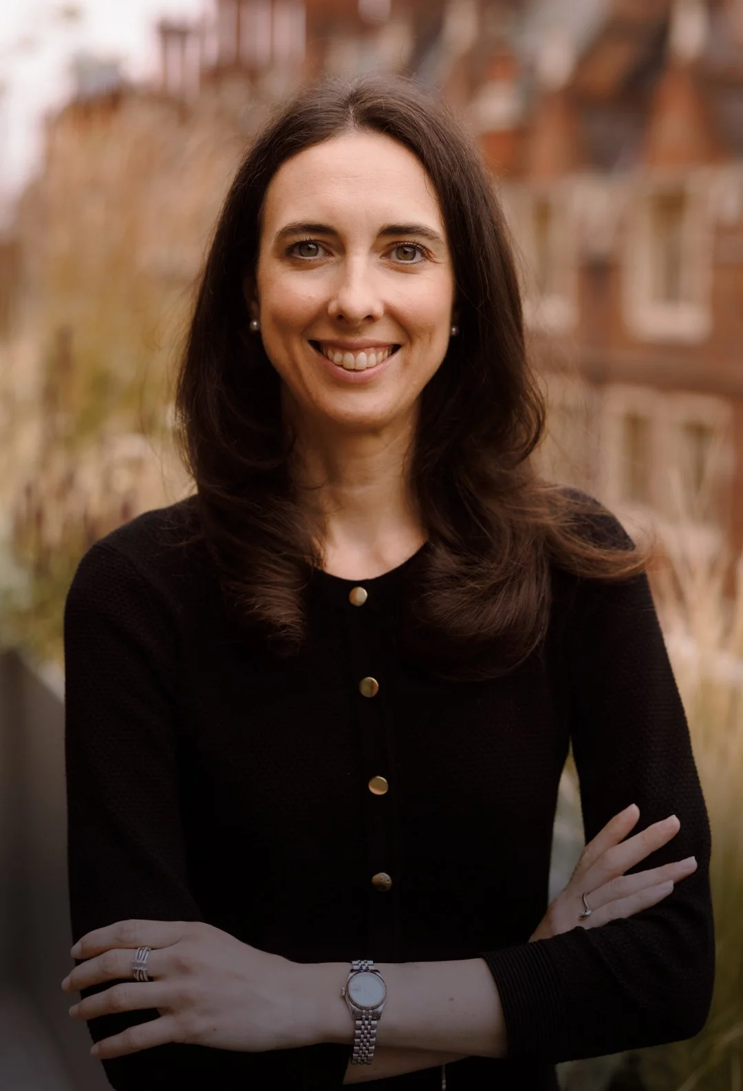 A smiling woman with dark brown hair, wearing a black top with gold buttons, standing outdoors with blurred buildings and trees in the background.