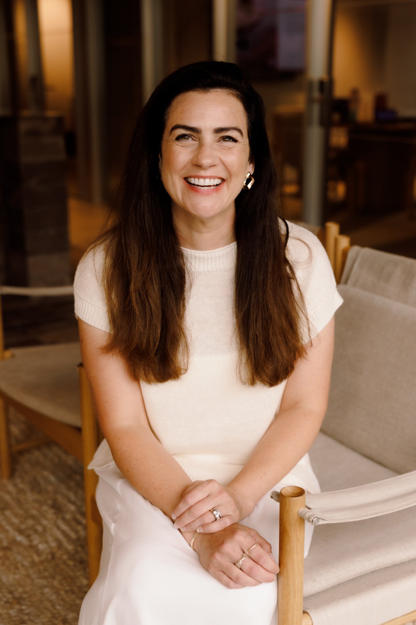 A woman with long dark hair, smiling, wearing a cream-colored top and earrings, sitting on a beige chair in a warmly lit room with wooden furniture.
