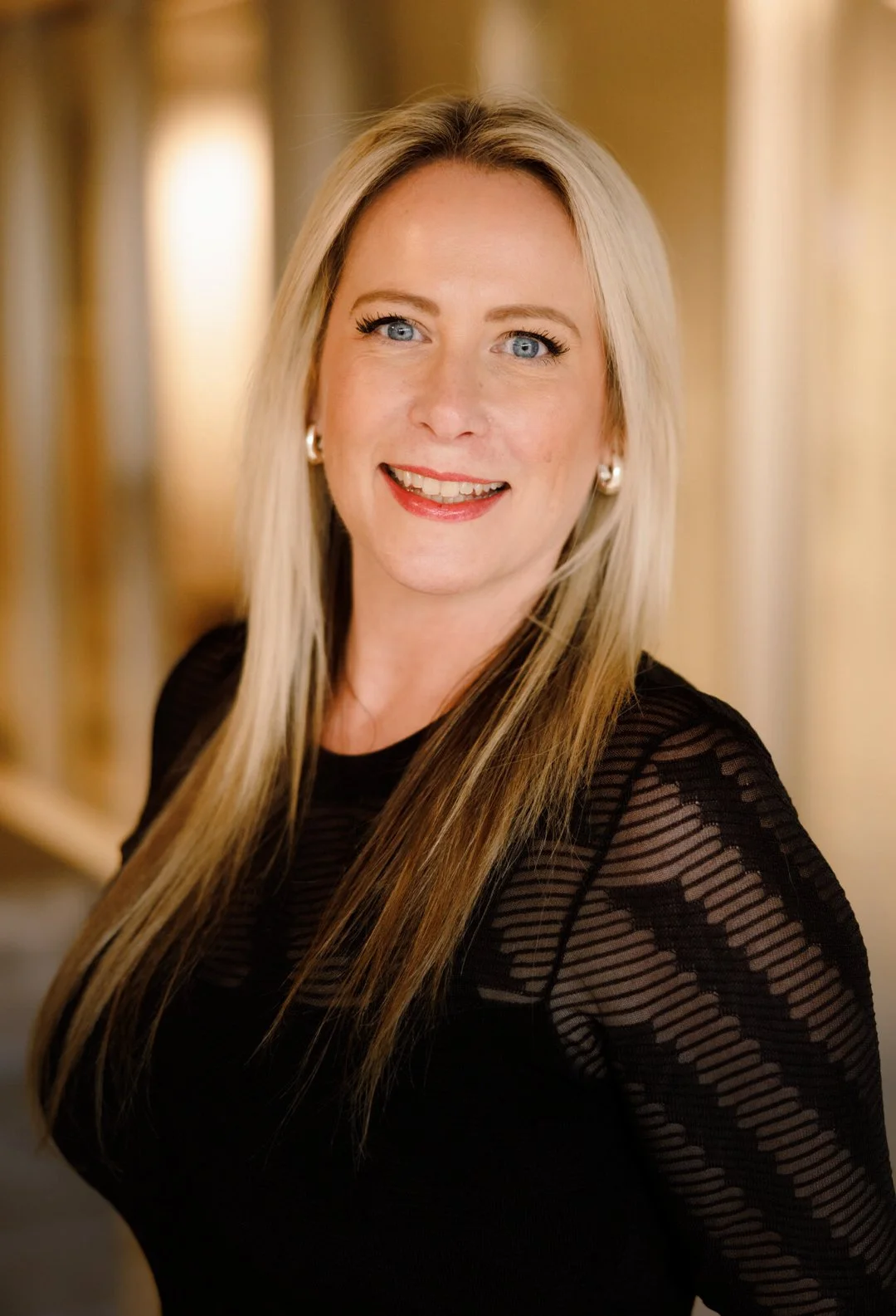 A woman with long blonde hair and blue eyes smiling at the camera, wearing earrings and a black top with sheer striped sleeves, in a warmly lit indoor setting.