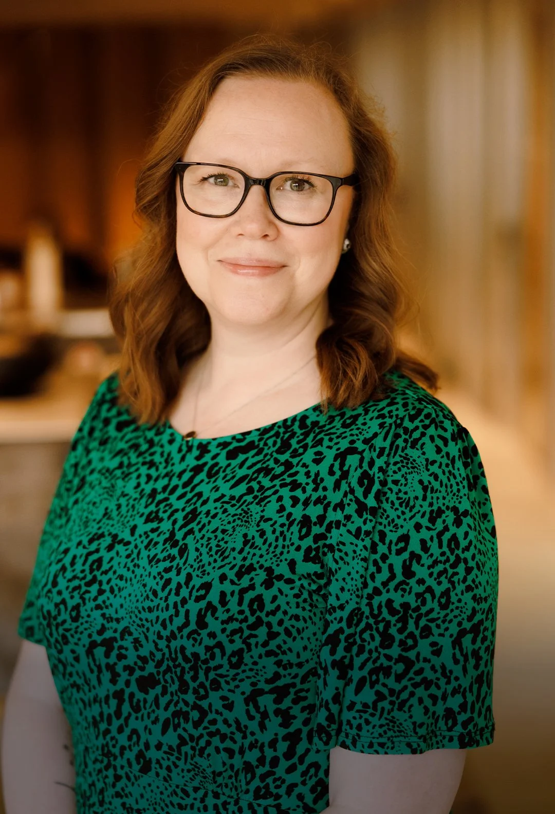 A woman with shoulder-length reddish-brown hair wearing glasses and a green dress with a black animal print pattern, standing indoors with a blurred background.