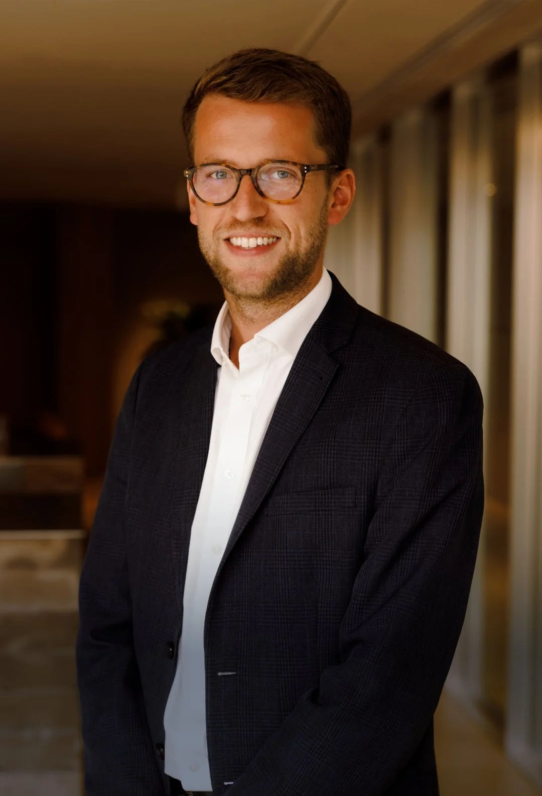 A young man with brown hair, glasses, and a beard, wearing a black suit jacket and white dress shirt, smiling in an indoor setting with blurred background.