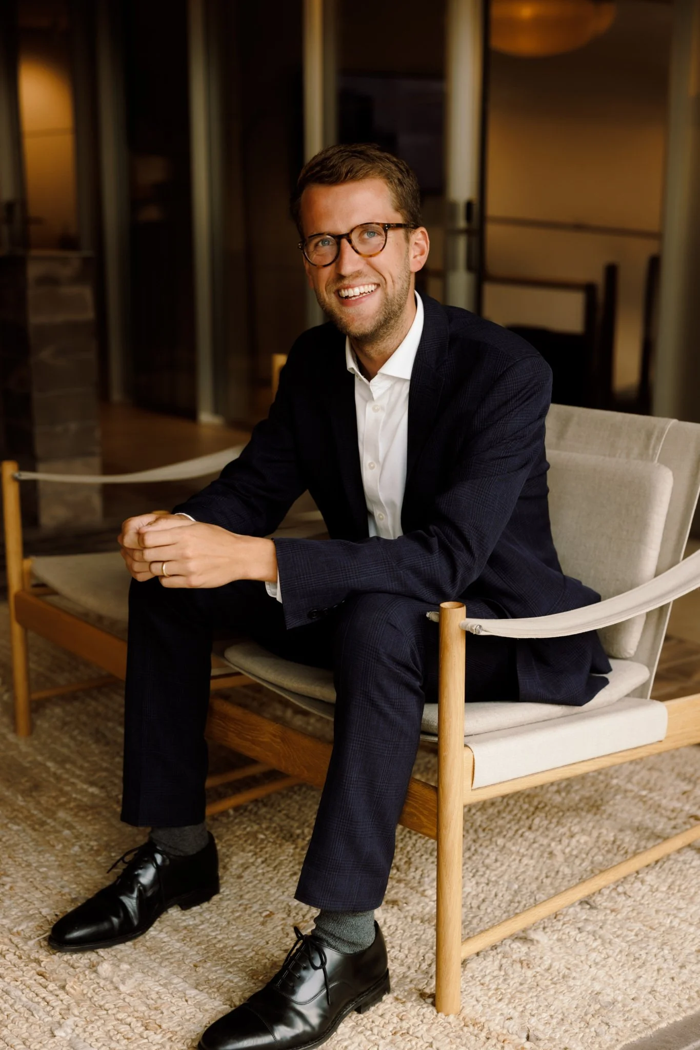A man wearing glasses, a white shirt, and a dark suit sitting on a wooden chair in a warmly lit room.