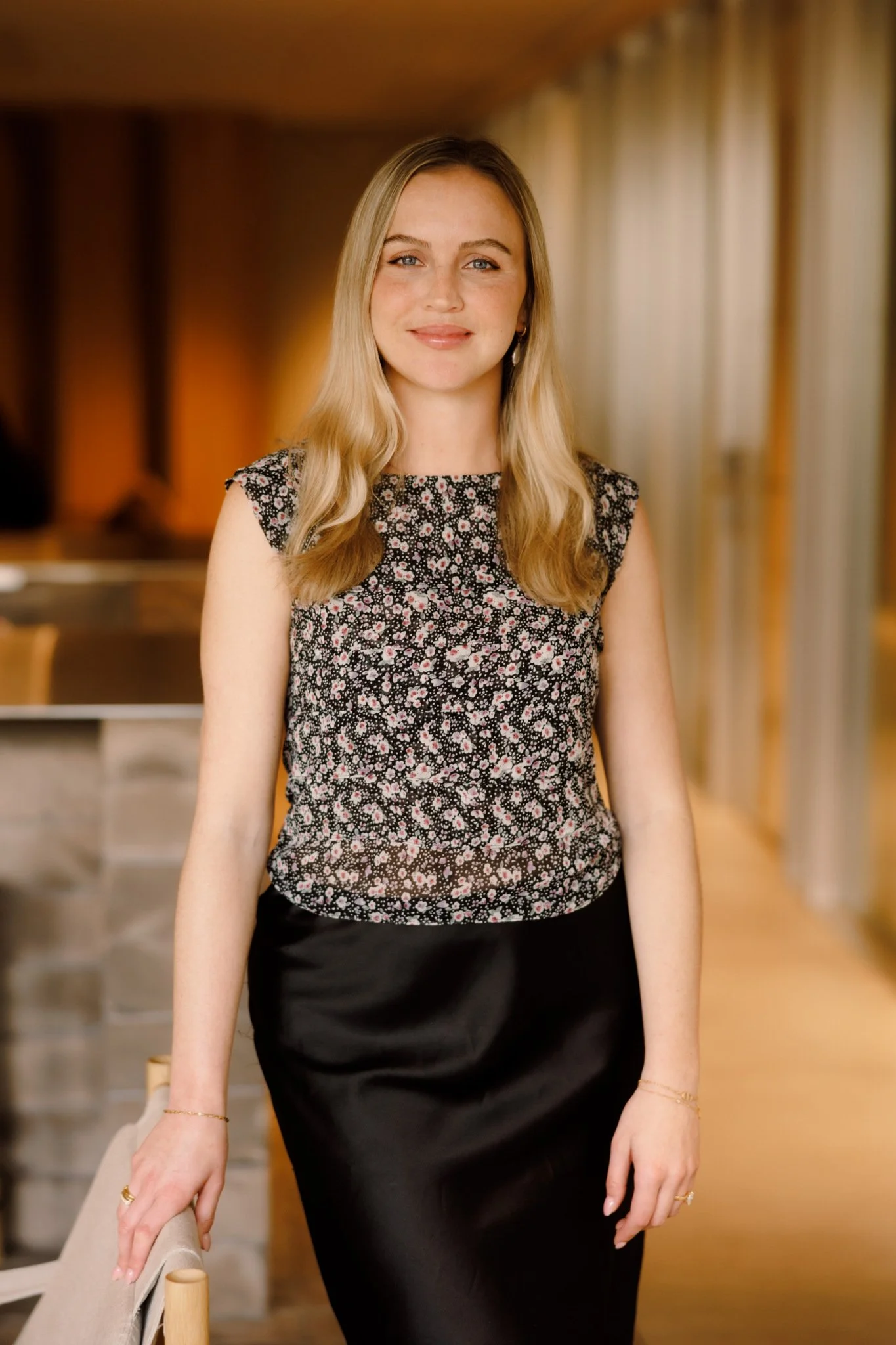 A woman with long blonde hair, wearing a black floral sleeveless top and a black skirt, standing in a warmly lit room with beige curtains in the background.