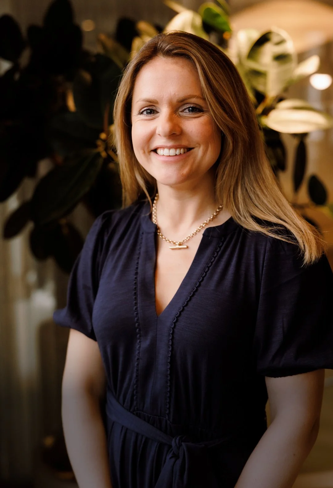 A woman with shoulder-length brown hair, wearing a black dress and a pearl necklace, standing indoors with a large plant in the background.