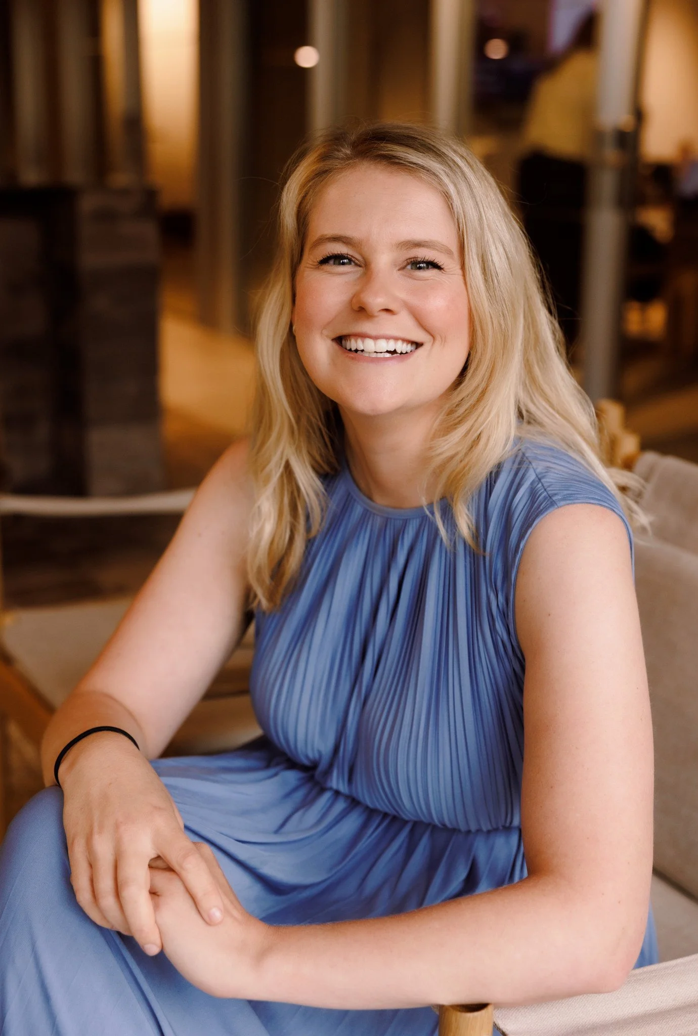 A young woman with blonde hair smiling, wearing a blue pleated dress, sitting indoors with a blurred background.