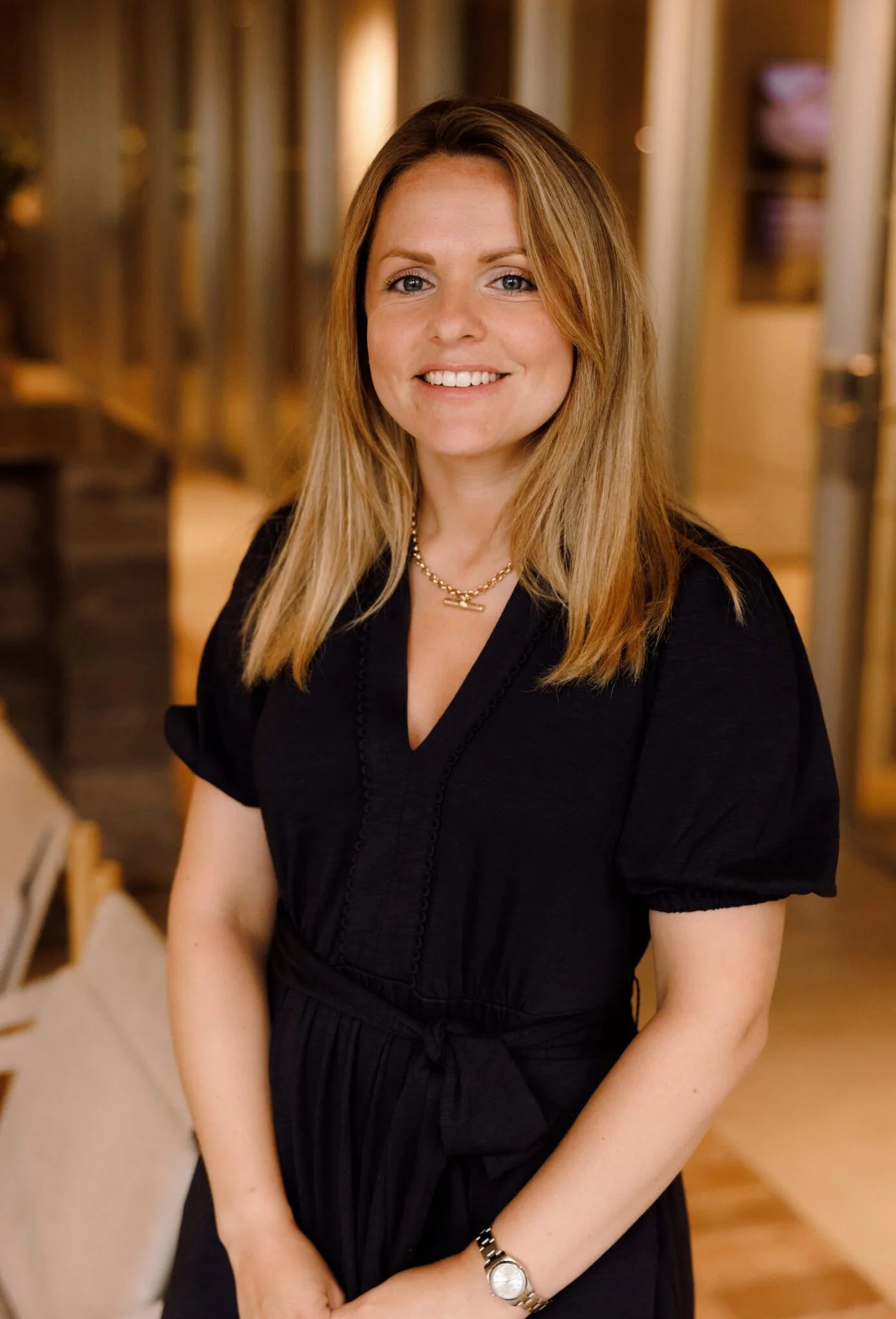 A smiling woman with long, light brown hair wearing a black dress and jewelry, standing indoors with wooden shelves and warm lighting in the background.