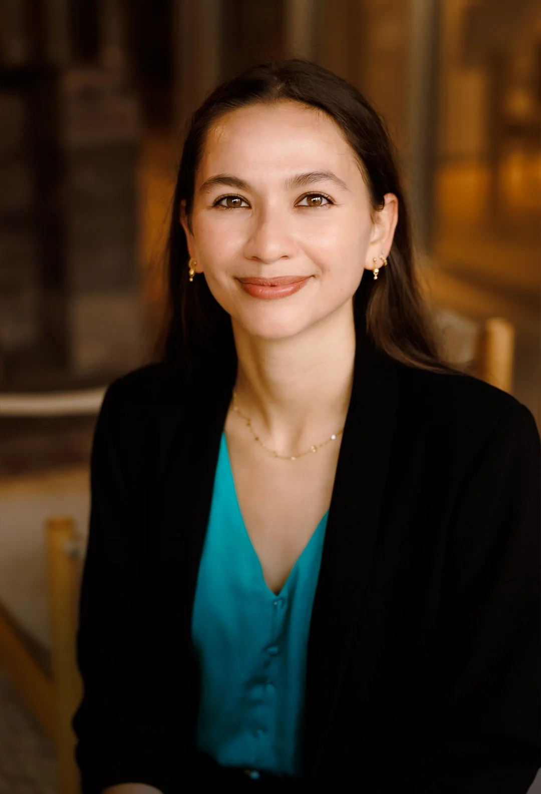 Portrait of a young woman with long brown hair, smiling, wearing gold earrings, a delicate necklace, a turquoise blouse, and a black blazer, indoors with warm lighting.