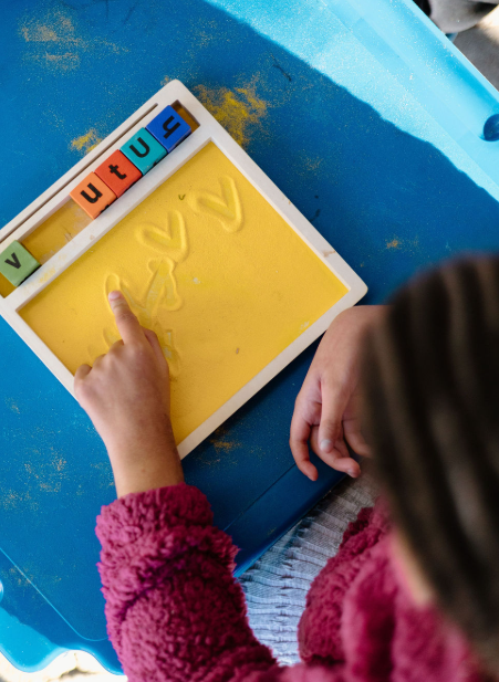 Child using a finger to trace letters in sand on a white tray with magnetic letters spelling 'V', 'U', 'T', 'Y', and '4' nearby.