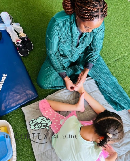 A woman performing a physical therapy exercise on a young girl lying on a mat, with exercise equipment nearby.