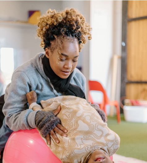 A woman leaning over a child lying on a pink exercise ball in a playroom.