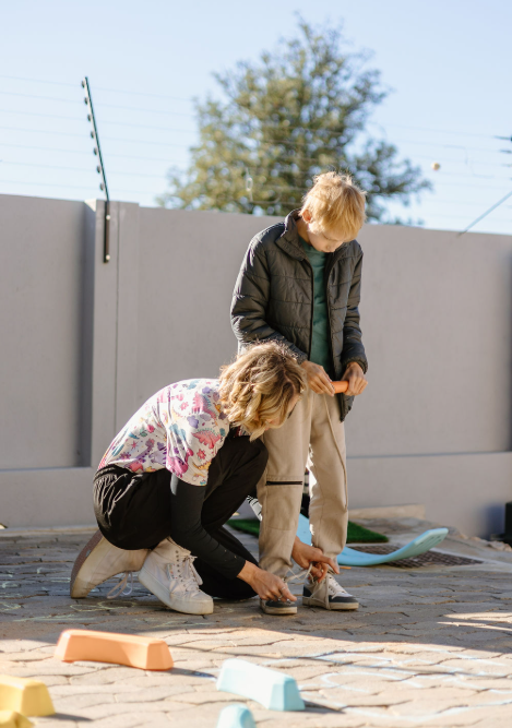 A woman helping a young boy tie his shoelaces outdoors on a stone pathway, with a gray fence and trees in the background.