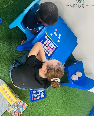 An adult and a child engaged in an educational activity at a blue desk with colorful tiles. The adult is laying on the floor, while the child is sitting at the desk. There are educational books and a water bottle nearby on a green carpeted floor.