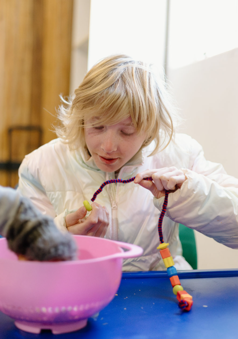 A young girl with blonde hair playing with a colorful beaded toy near a pink bowl, in a bright room with natural light.