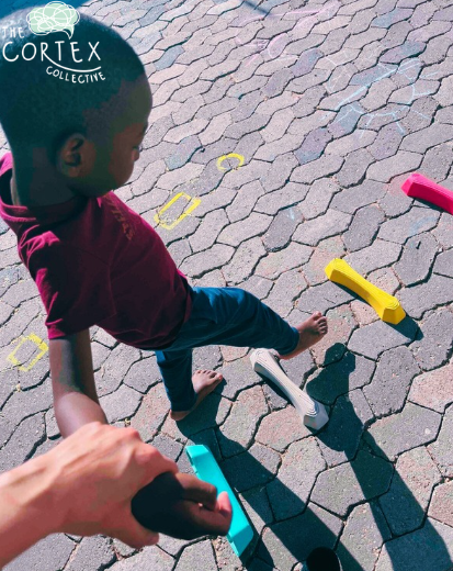 Child standing on a paved area with colorful toy cars and yellow chalk markings, with a person's hand holding a camera or phone for a selfie.