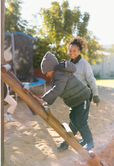 A young child in a gray hat and jacket climbing a slide while an adult woman watches and smiles outdoors on a sunny day.
