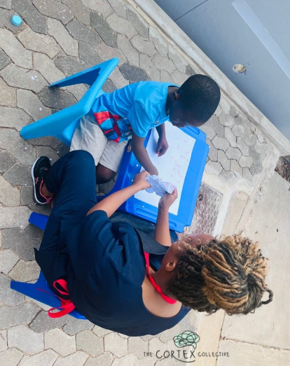 A young girl with blonde braids sitting on the ground playing with a blue whiteboard and some papers, while a boy with dark hair standing next to her looks at the whiteboard, outdoors on a paved surface.