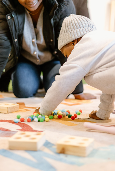 Child in a gray beanie and white hoodie playing with colorful pom-poms on a rug, with an adult beside him.