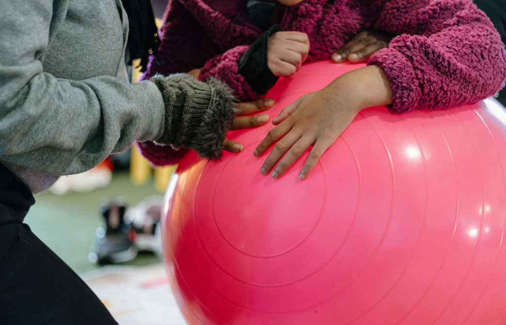 Two children hold hands and rest their hands on a large pink exercise ball in a gym or fitness setting.