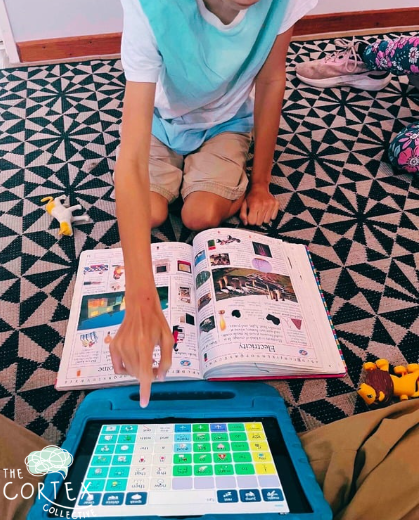 Child sitting on a patterned carpet, pointing at a tablet with a coloring app, with an open magazine or catalog in front of them.