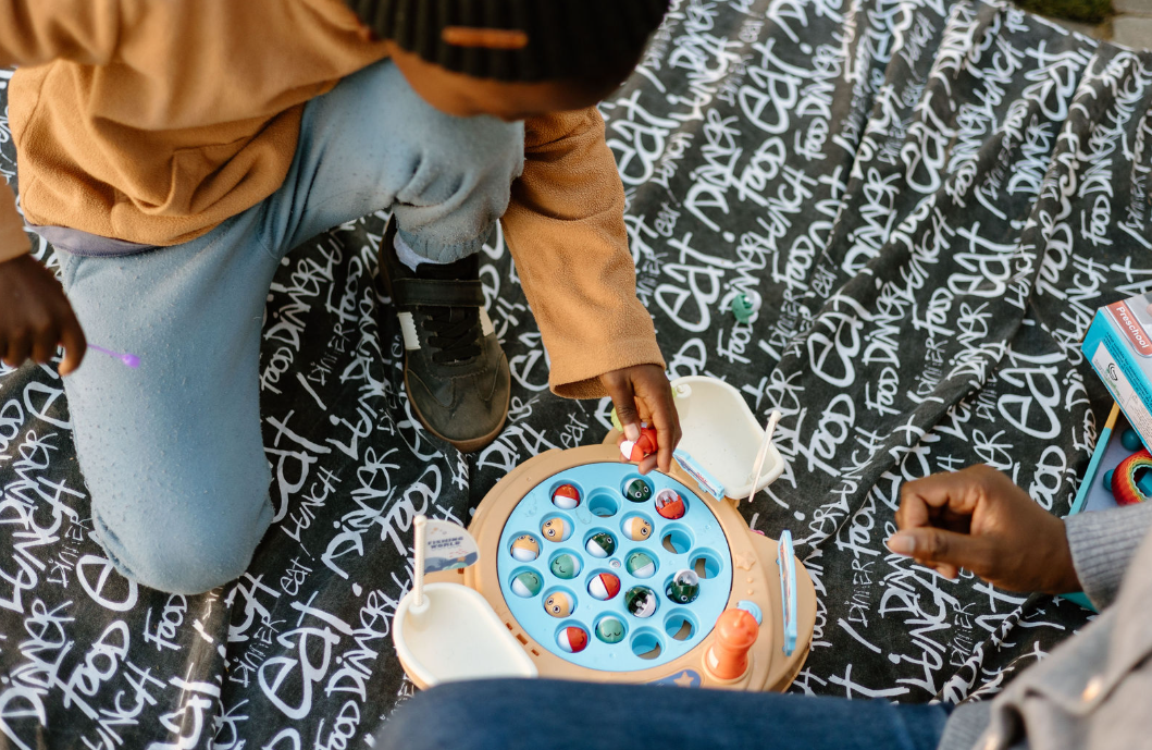 Two children playing a fishing game on a black and white cloth with graffiti-style text. One child is reaching for the game pieces while the other is sitting nearby.