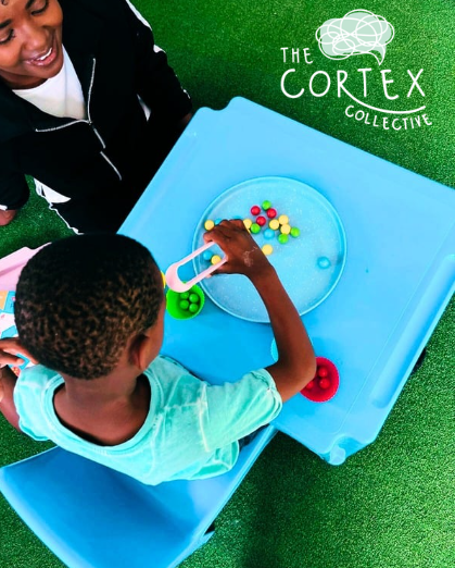 A young boy playing a game of bowling with colorful balls in a clear bowl, while an adult watches. They are at a blue table outside on green artificial turf, with the logo of The Cortex Collective in the top right corner.