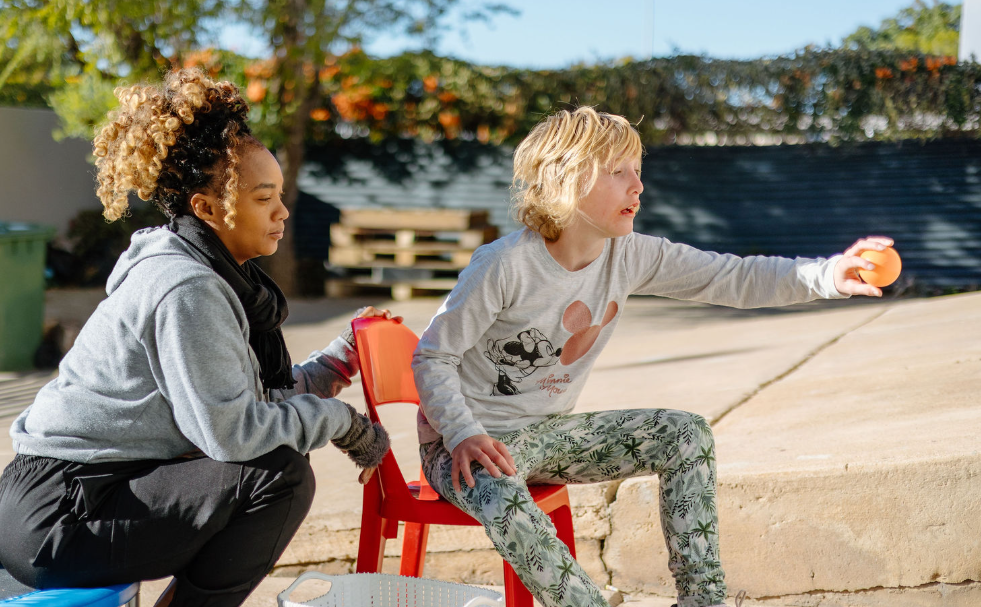 A young girl with blonde hair sitting on a red chair, reaching out to throw a small orange ball, while a young woman with curly hair squats beside her, supporting her.