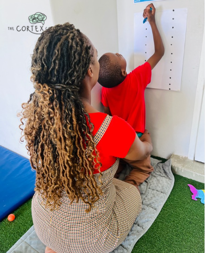 A woman with long, curly hair sitting on a cushioned mat, watching a boy in a red shirt write on a paper attached to the wall.