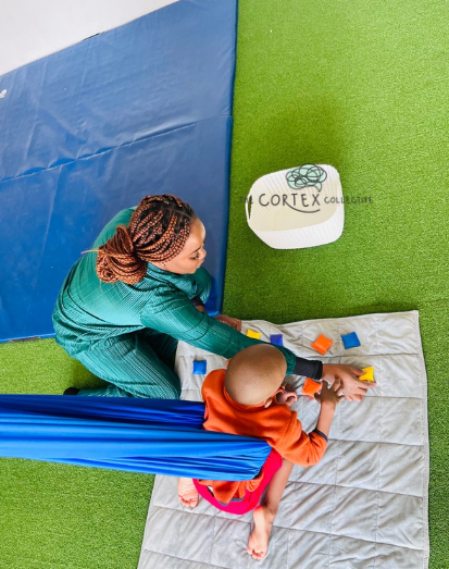 An adult woman with braided hair is assisting a child in an orange shirt with a brain injury in a wheelchair, with colorful blocks on a gray mat in a therapy room with green and blue walls.