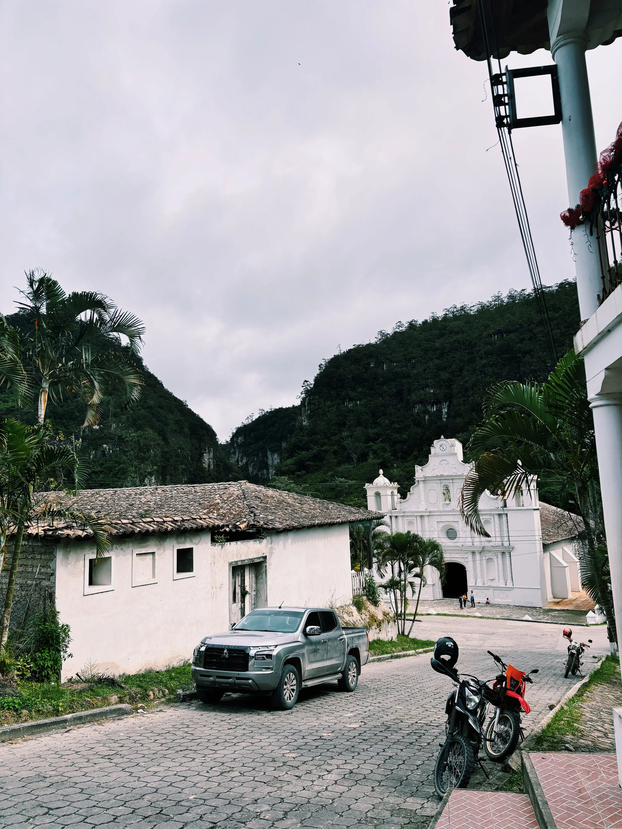 Vista de una calle empedrada con un coche gris, una moto y una bicicleta, una iglesia blanca al fondo y montañas cubiertas de vegetación al fondo, cielo nublado.