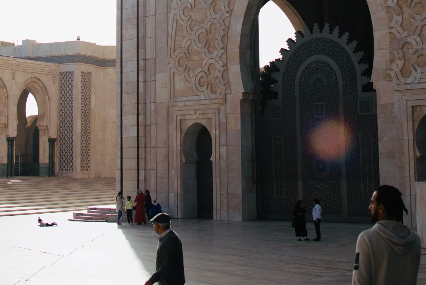 Personas caminando frente a una gran estructura con arquitectura islámica, arcos y puertas decoradas, en una explanada con escaleras.