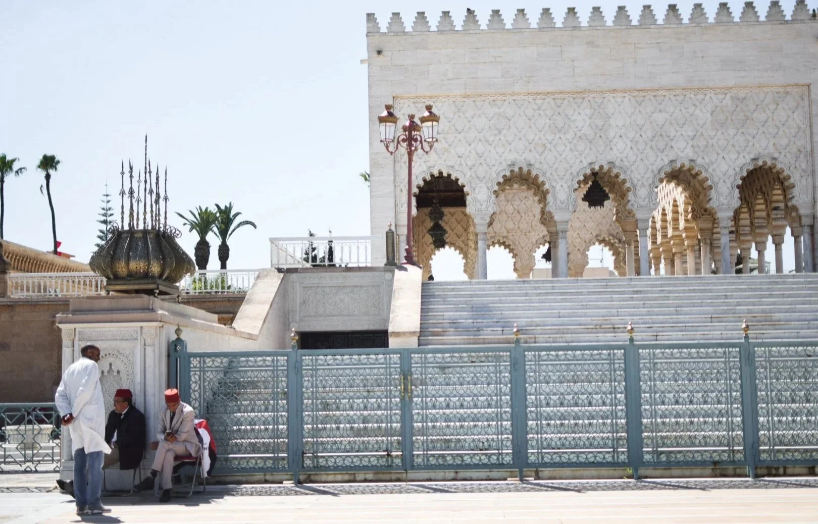 Imagen del exterior de una mezquita, con escalones de mármol y columnas con arcos. Cuatro hombres con vestimenta tradicional, uno de pie y tres sentados en una banca, en primer plano. Palmas en el fondo.