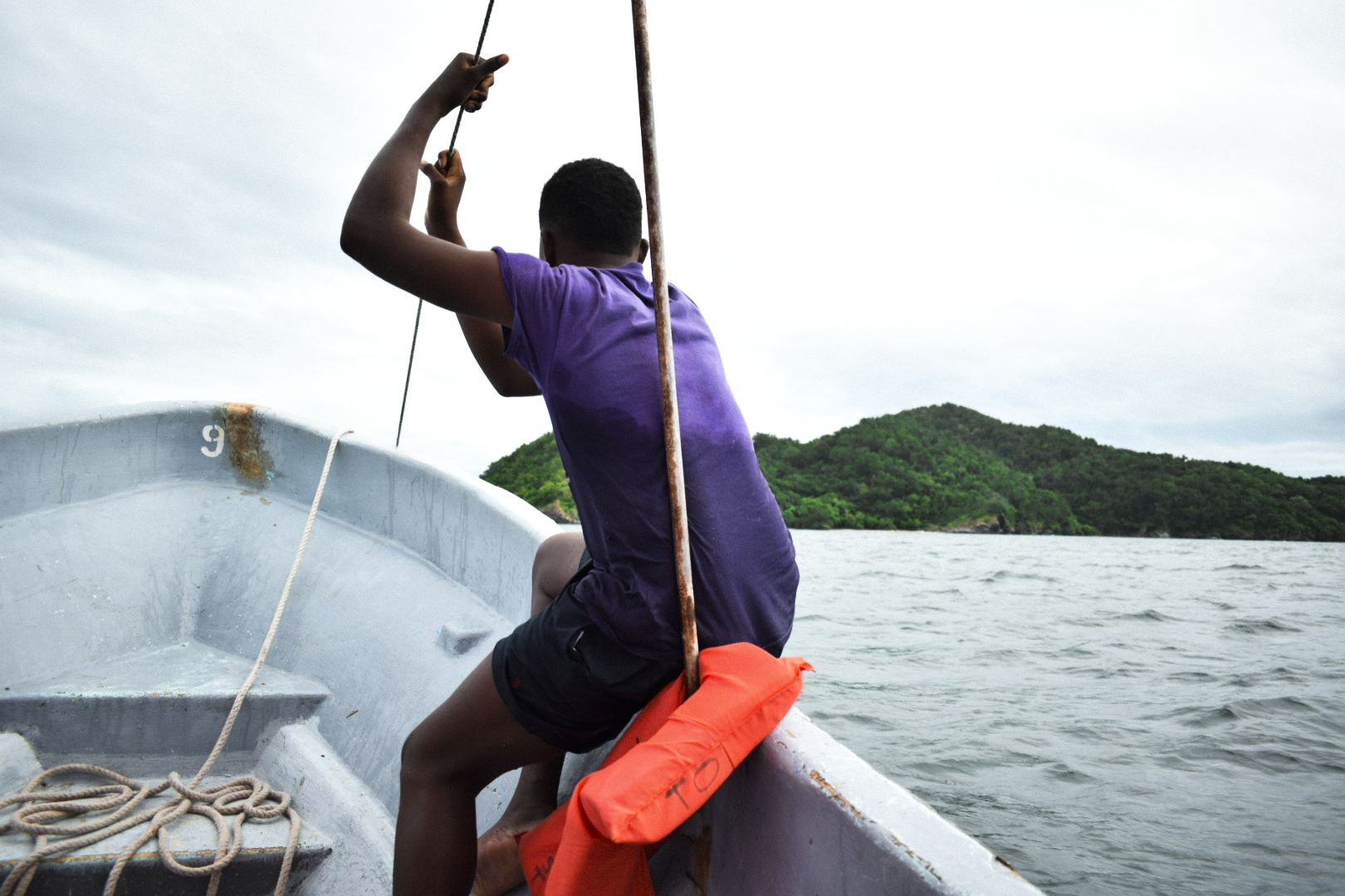 Joven en un bote de madera remando en el agua con una lancha y un paisaje de islas verdes de fondo.