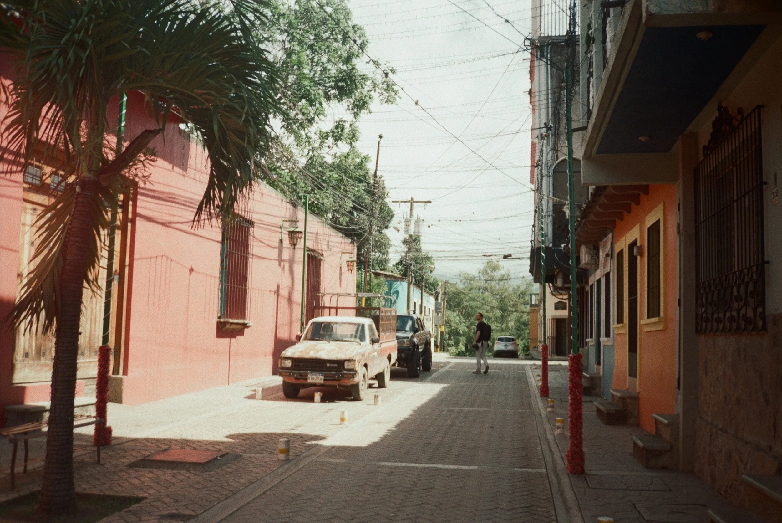 Calle tranquila en un pueblo, con casas coloridas, autos estacionados, un hombre caminando y árboles en el fondo.