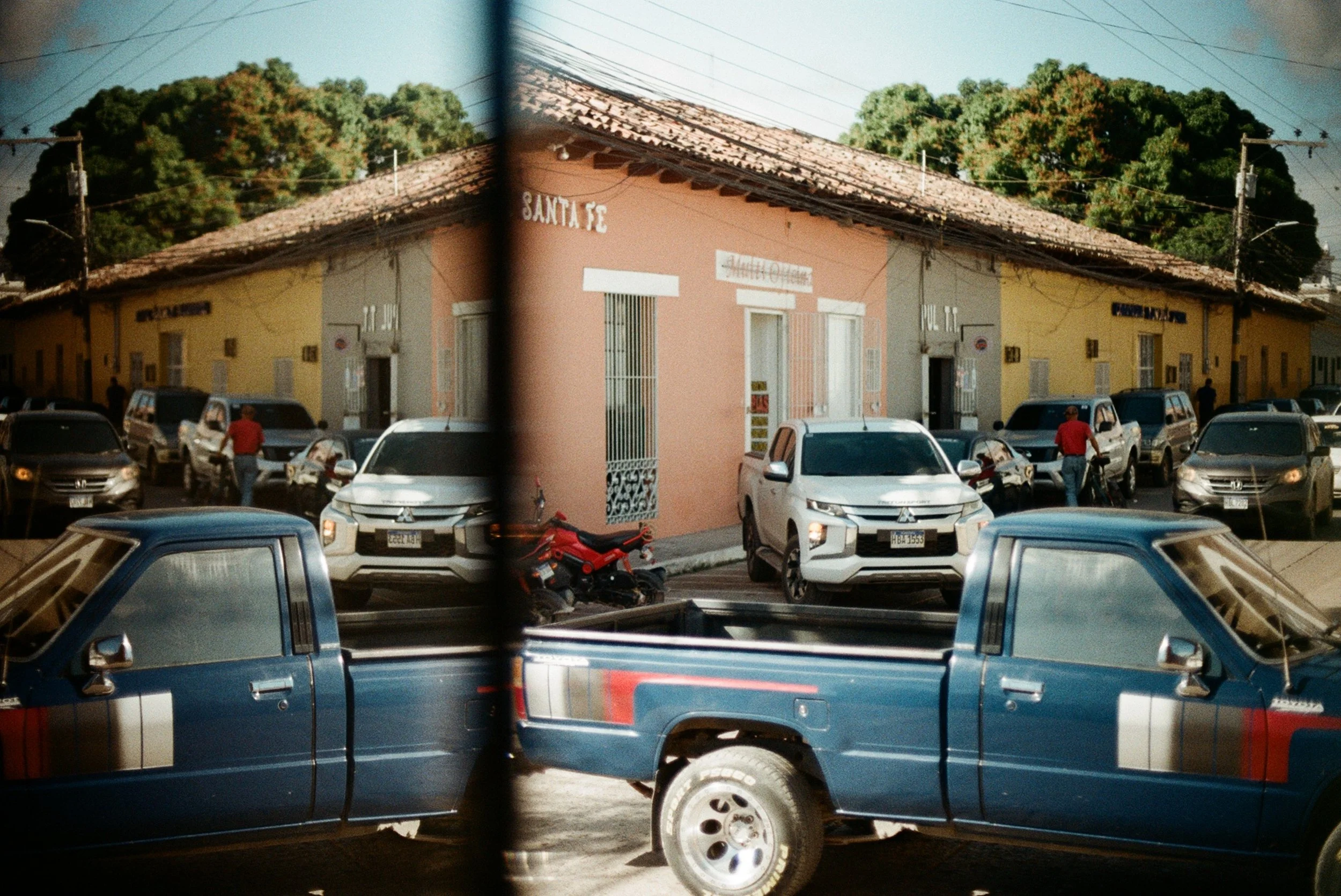 Vista de un estacionamiento con varios autos y un edificio de color rosado con ventanas con rejas, en una zona con árboles verdes al fondo, y un espejo que refleja parte de la escena.