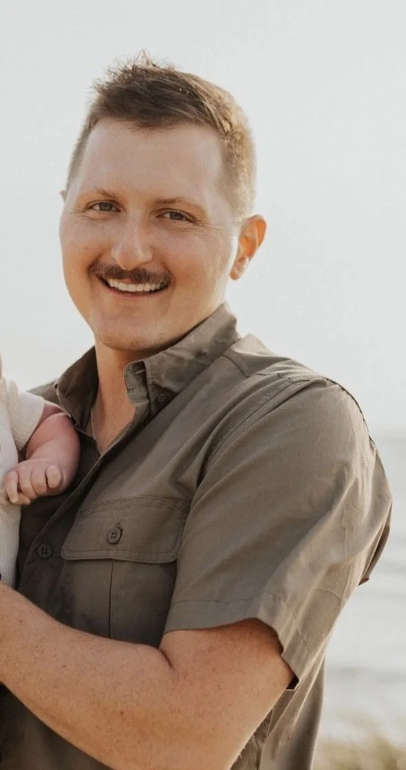 A man with short hair, a mustache, smiling, wearing a short-sleeved button-up shirt, holding a baby.