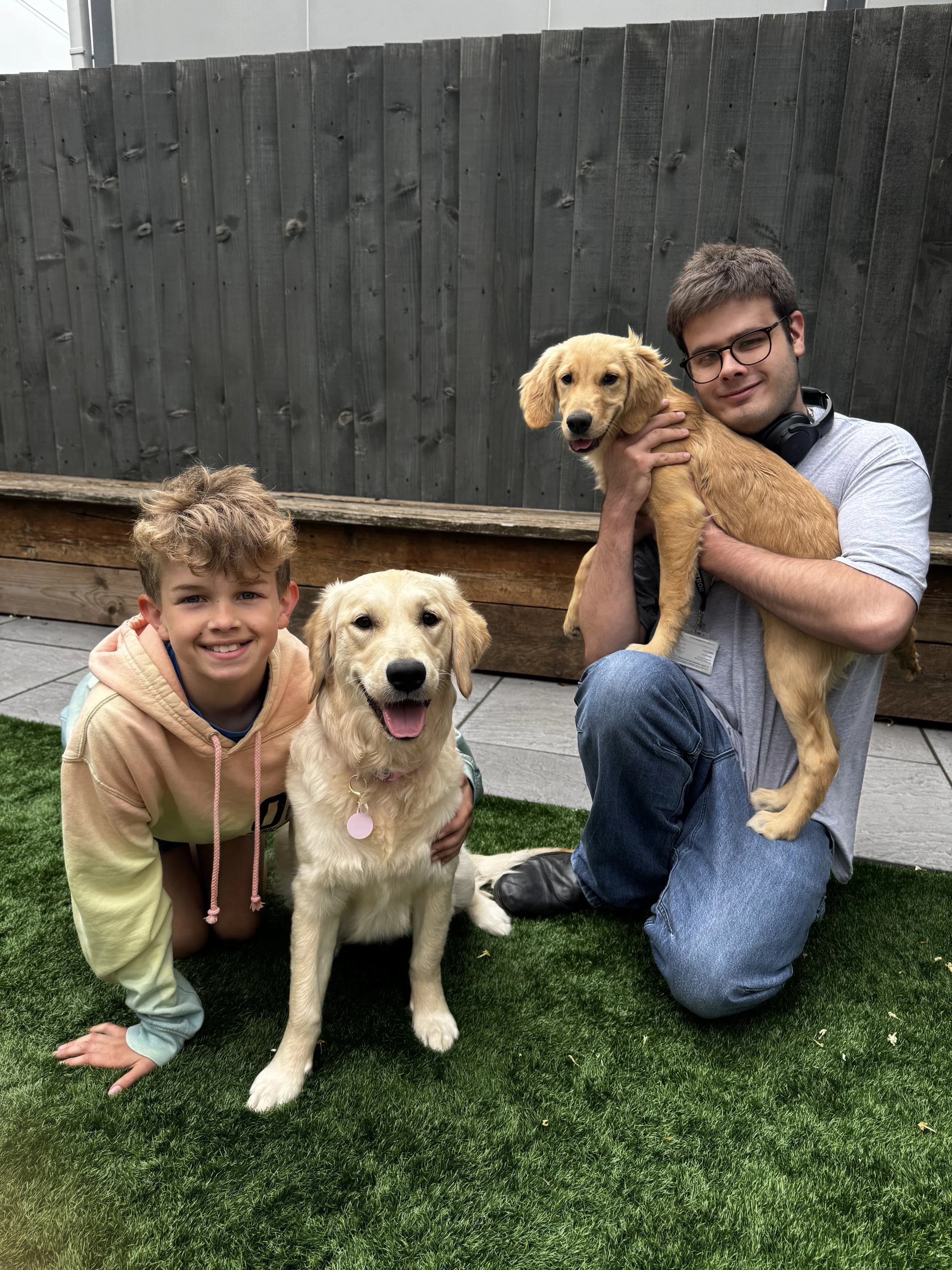 Two children and two golden retrievers in a backyard with a wooden fence. One child is kneeling with a dog, the other is holding a dog, all smiling.
