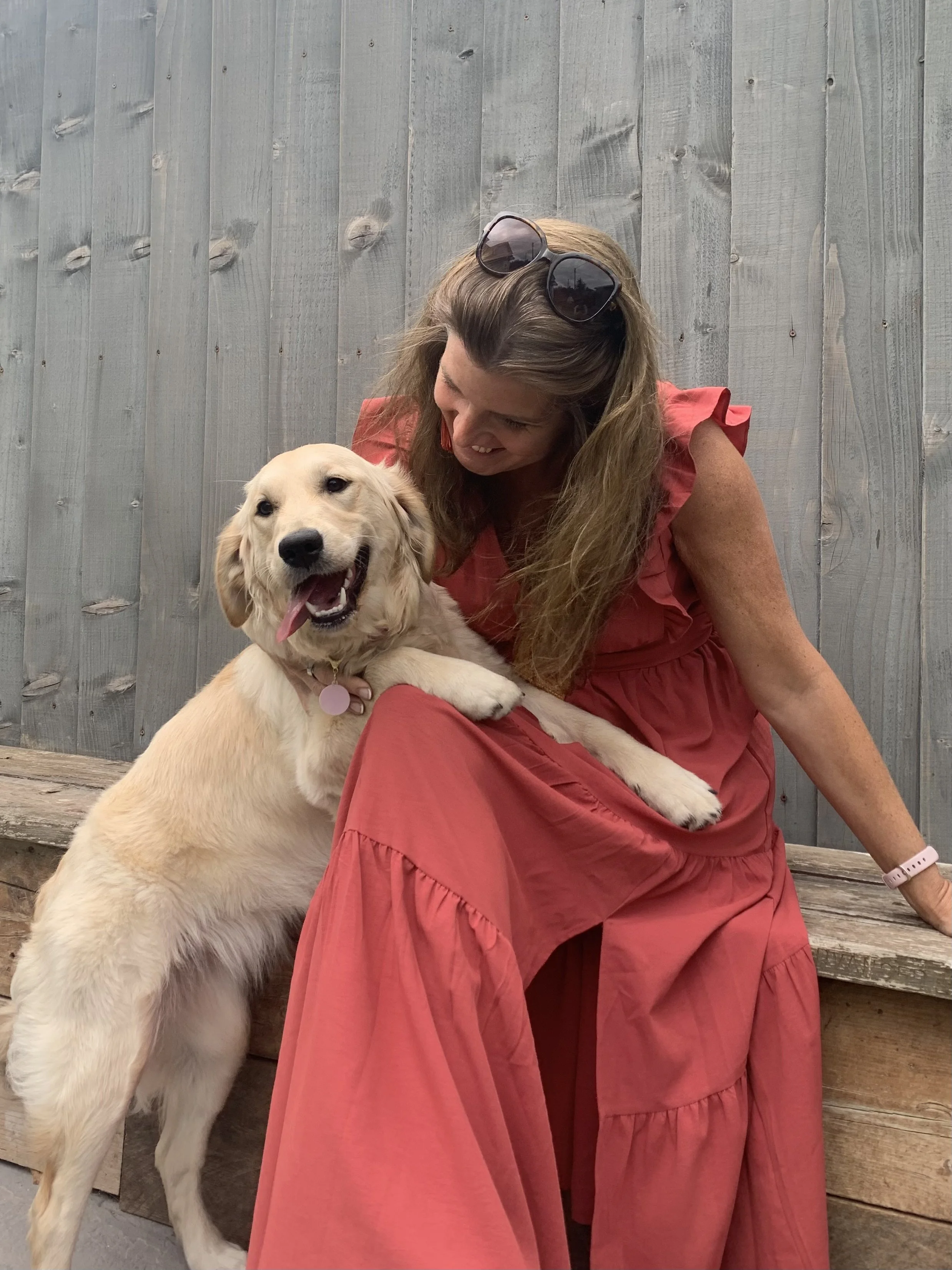 A woman in a long red dress sitting on a wooden bench, smiling as a Golden Retriever puppy leans on her lap and playfully licks her.