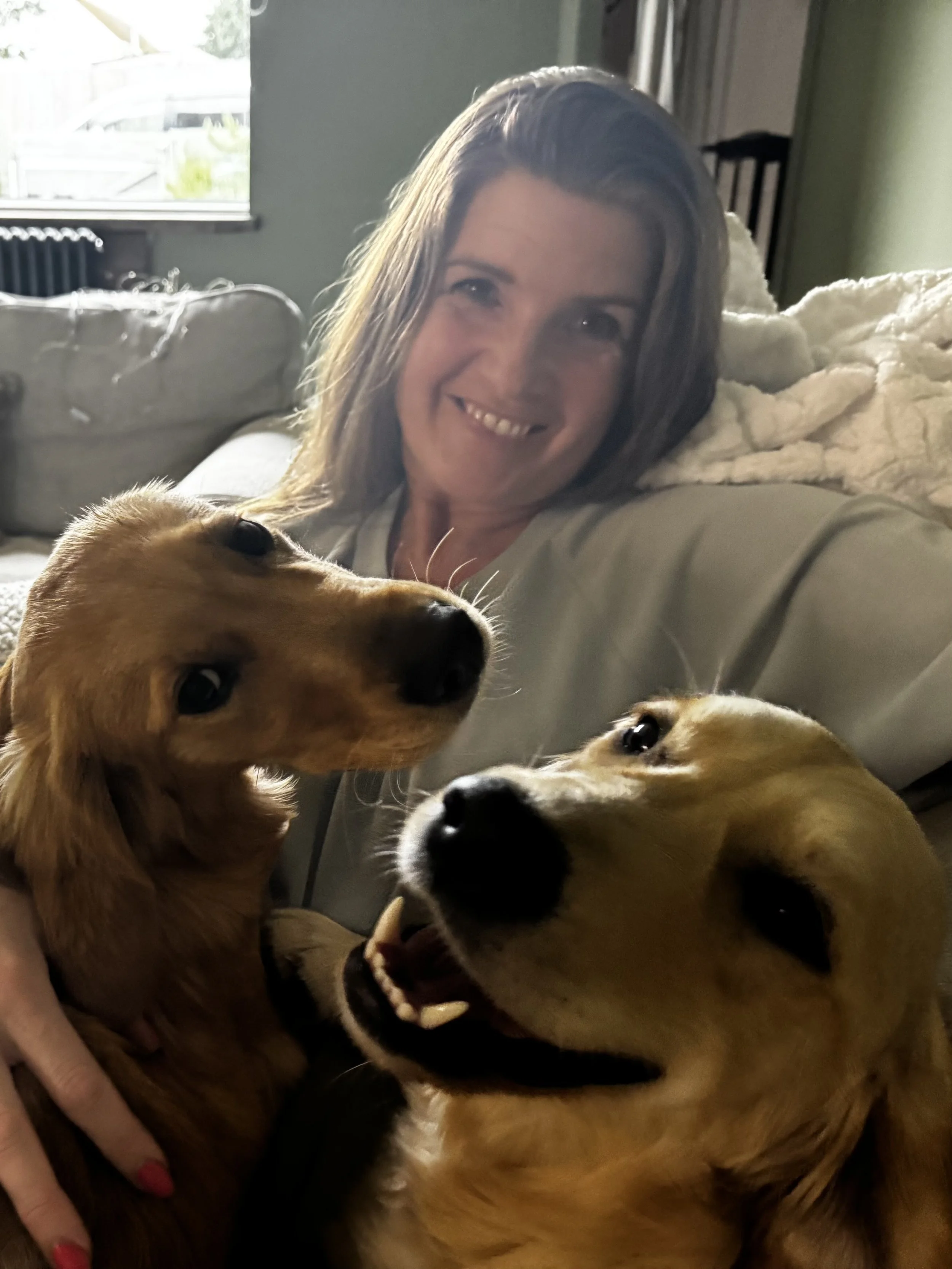 Smiling woman with brown hair on a couch, holding two golden retrievers, one with a gentle expression and the other with a playful, open-mouthed look.