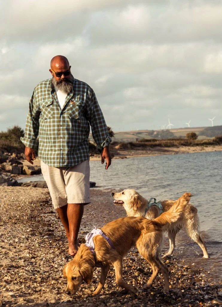 A man with a beard and sunglasses walking barefoot along a rocky beach with two golden retriever dogs.