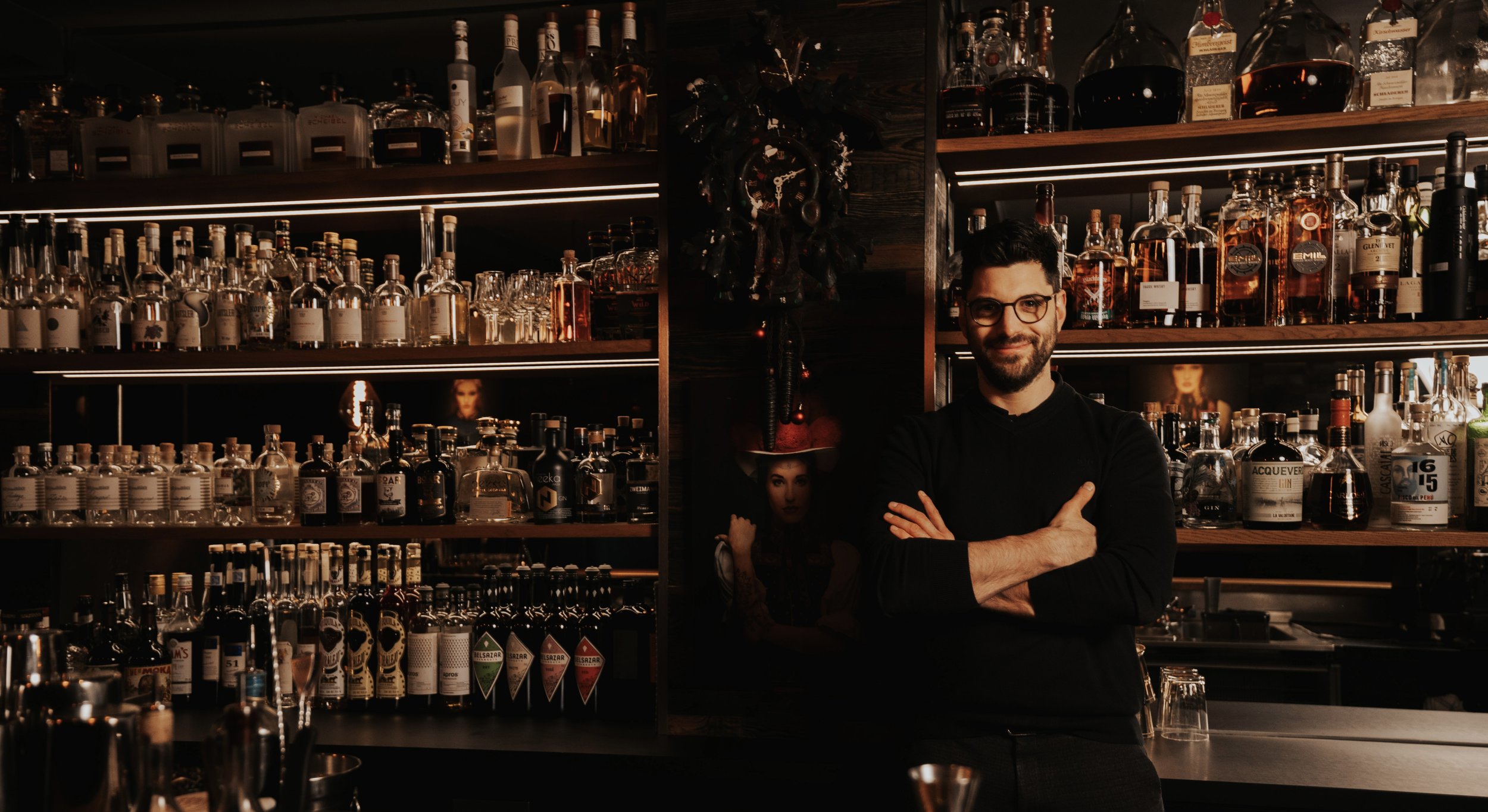 Cocktailkurs in Hamburg. Ein Barkeeper steht in einer Bar vor einer Wand mit verschiedenen Flaschen und Gläsern, trägt eine schwarze Uniform, trägt Brille und lächelt.