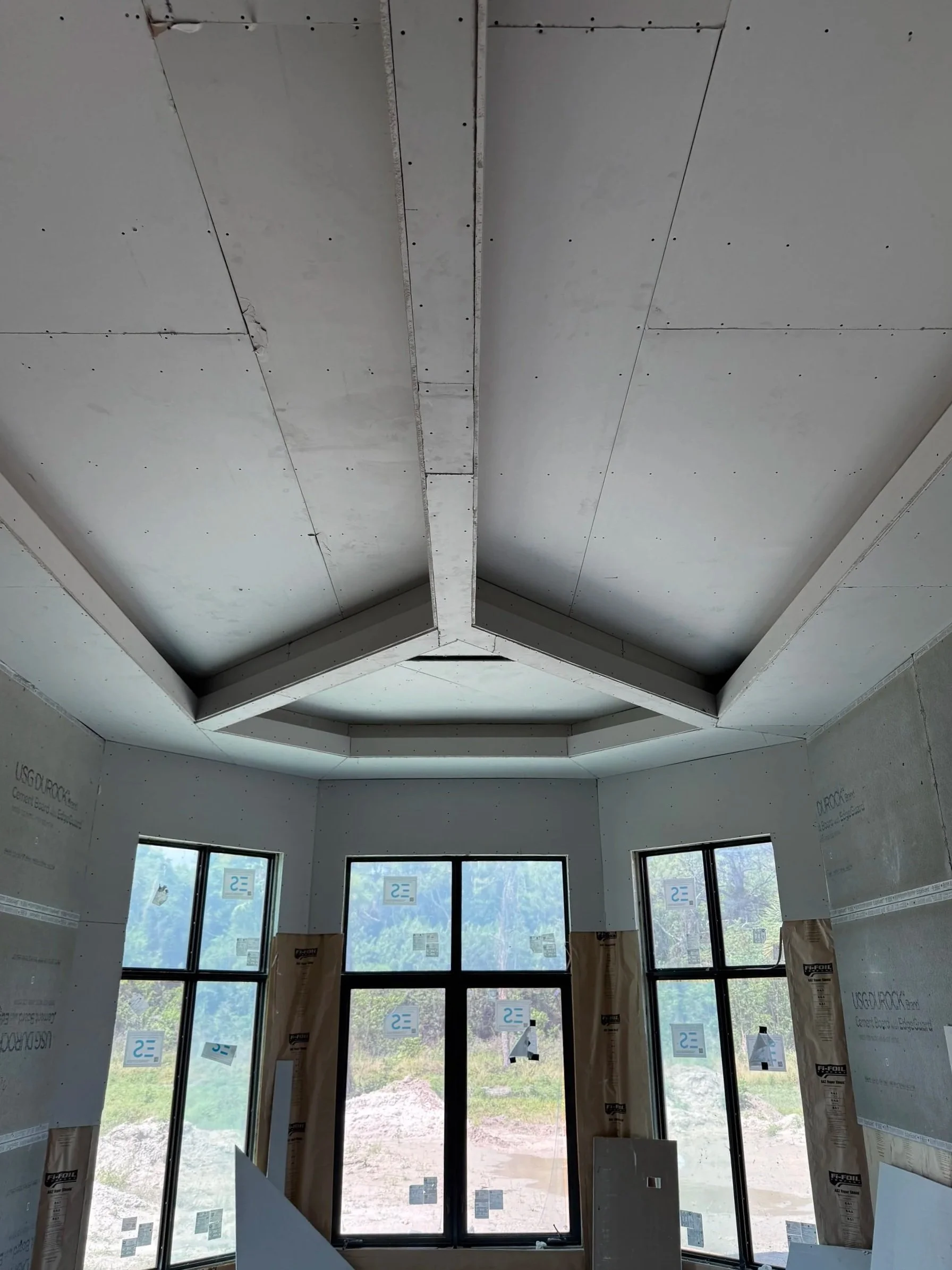 Interior of a room under construction with drywall ceiling and big windows showing an outdoor landscape.