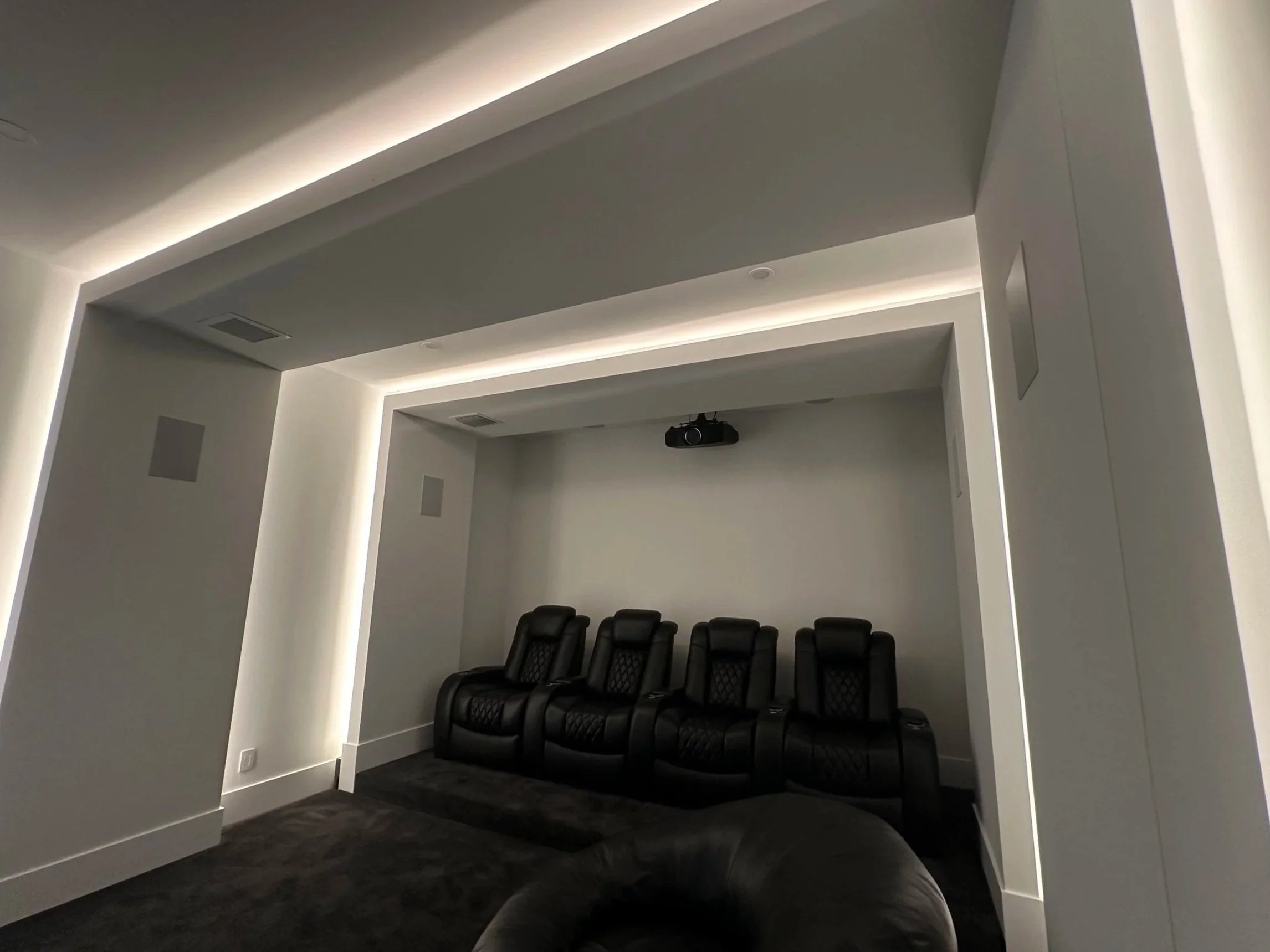Home theater room with five black leather chairs, a projector mounted on the ceiling, and adjustable lighting along the ceiling edges.
