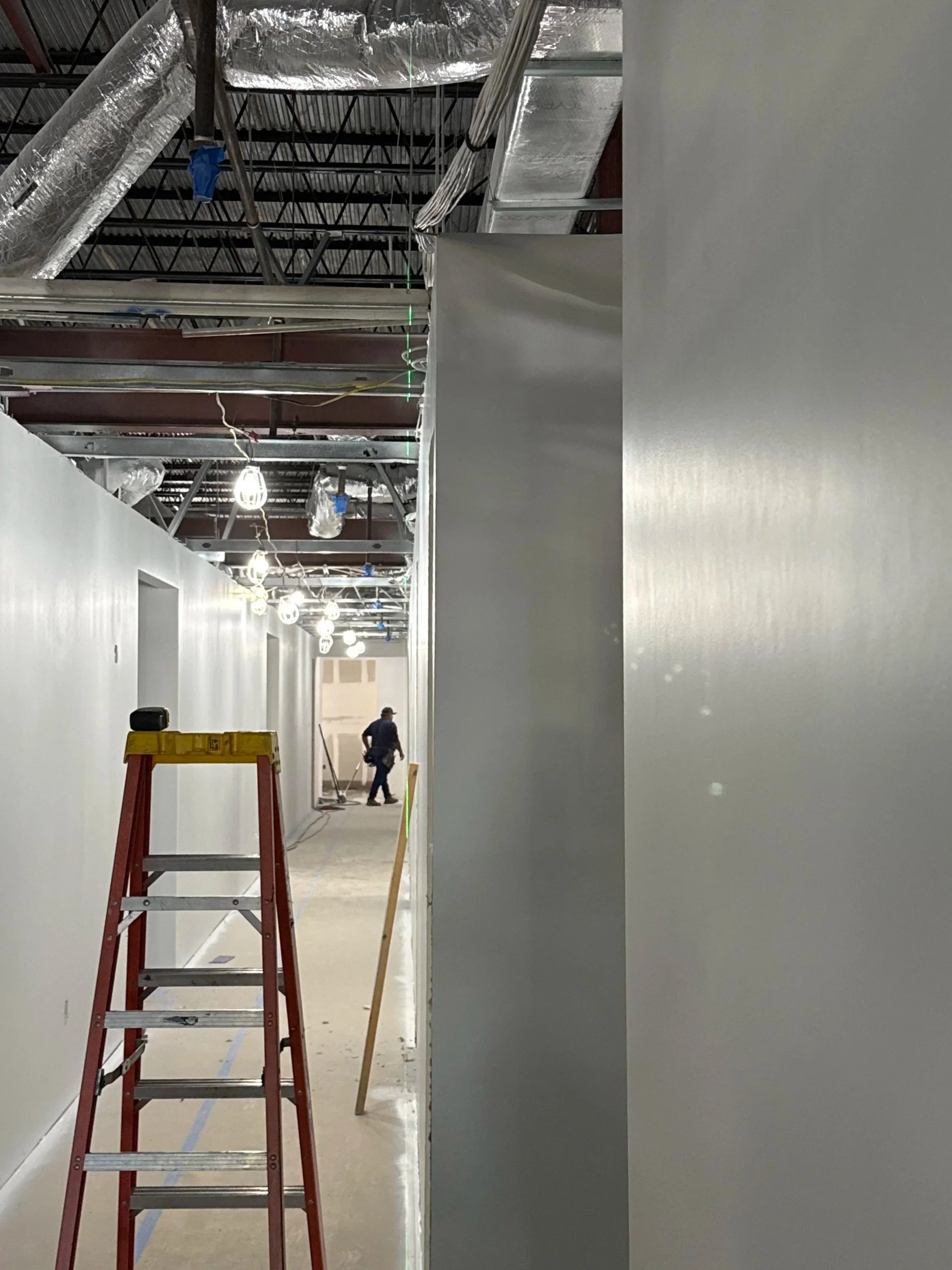 Interior hallway under construction with a red ladder, construction worker, and exposed ceiling infrastructure.