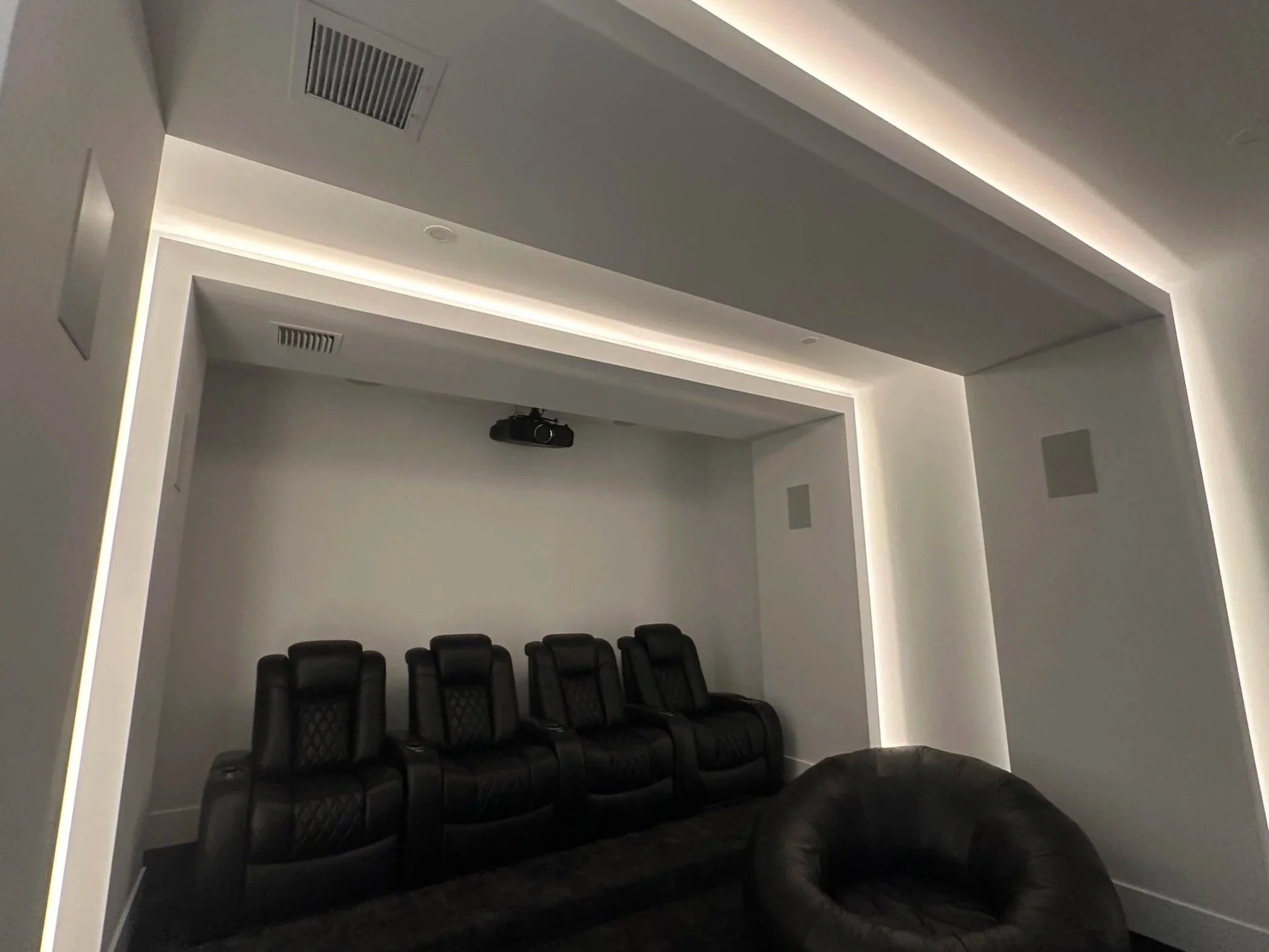 A modern home theater room with five black leather recliner chairs in a row, a black leather bean bag at the front, a ceiling-mounted projector, and a white ceiling with recessed lighting and air vents.